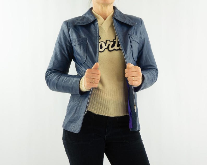 K&ouml;nnte beinhalten: Blaue Lederjacke mit Kragen und Rei&szlig;verschluss. Die Jacke hat zwei Vordertaschen und wird &uuml;ber einem beigen Pullover getragen. Die Person tr&auml;gt auch eine schwarze Hose.