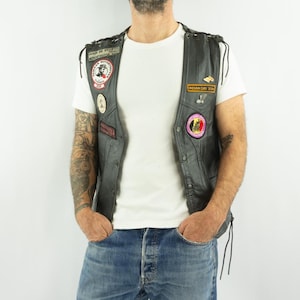 Peut inclure: Gilet en cuir noir avec plusieurs écussons, dont un écusson circulaire avec un motif rouge et blanc. Le gilet a une fermeture boutonnée et des côtés lacés. La personne porte un t-shirt blanc et un jean bleu.