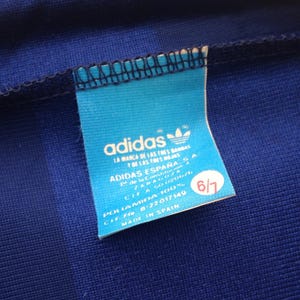 K&ouml;nnte beinhalten: Ein blaues Kleidungsst&uuml;ck aus Stoff mit einem hellblauen rechteckigen Etikett, das das adidas-Logo und Text wie ADIDAS ESPA&Ntilde;A S.A., POLIAMIDA 100%, MADE IN SPAIN sowie einen roten runden Aufkleber mit der wei&szlig;en Zahl 6/7 aufweist.
