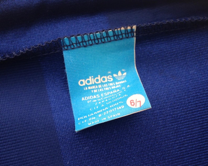 K&ouml;nnte beinhalten: Ein blaues Kleidungsst&uuml;ck aus Stoff mit einem hellblauen rechteckigen Etikett, das das adidas-Logo und Text wie ADIDAS ESPA&Ntilde;A S.A., POLIAMIDA 100%, MADE IN SPAIN sowie einen roten runden Aufkleber mit der wei&szlig;en Zahl 6/7 aufweist.