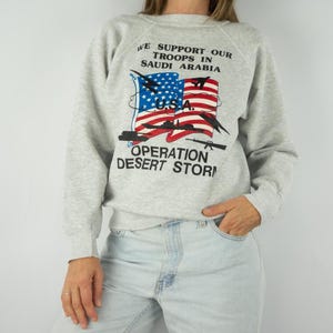May include: A light gray sweatshirt with the text "WE SUPPORT OUR TROOPS IN SAUDI ARABIA" above a graphic of the American flag and the words "OPERATION DESERT STORM". The sweatshirt has long sleeves.