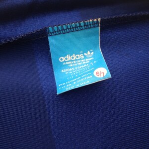 K&ouml;nnte beinhalten: Ein blaues Adidas-Etikett mit wei&szlig;em Text und einem Logo. Das Etikett lautet "adidas", "La Marca de las Tres Bandas", "Adidas Espana", "Polamida 100%", "CIF 8-22017149", "Made in Spain" und "6/7".
