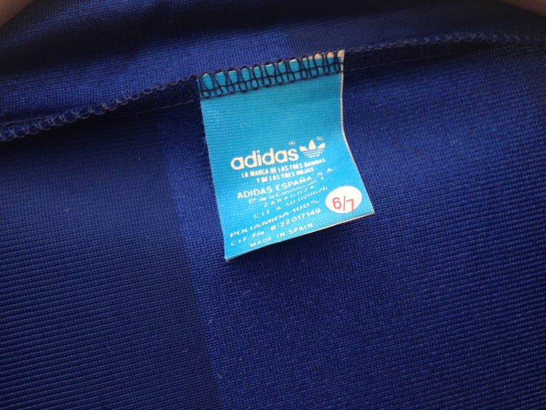 K&ouml;nnte beinhalten: Ein blaues Adidas-Etikett mit wei&szlig;em Text und einem Logo. Das Etikett lautet "adidas", "La Marca de las Tres Bandas", "Adidas Espana", "Polamida 100%", "CIF 8-22017149", "Made in Spain" und "6/7".