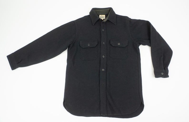 May include: A black long-sleeve shirt with two chest pockets and a button-down front. The shirt has a collared neckline and buttoned cuffs. The fabric appears to be a heavy, textured material, suitable for cooler weather.