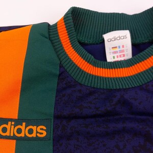 May include: Close-up of a vintage Adidas athletic shirt. The shirt is navy blue with orange and green accents. The collar and side panel feature orange stripes. The Adidas logo is prominently displayed in orange.