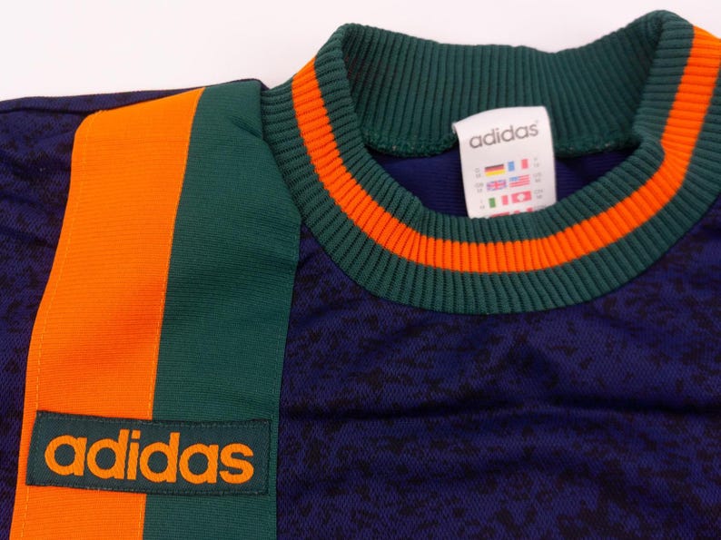 May include: Close-up of a vintage Adidas athletic shirt. The shirt is navy blue with orange and green accents. The collar and side panel feature orange stripes. The Adidas logo is prominently displayed in orange.