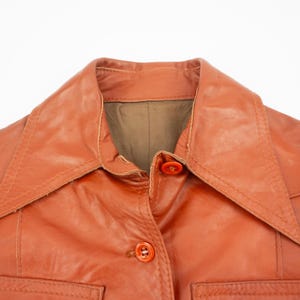 May include: Close-up of a vintage orange leather jacket. The jacket features a wide collar, two front pockets, and orange buttons. The leather has a slightly worn appearance, suggesting its age and use.