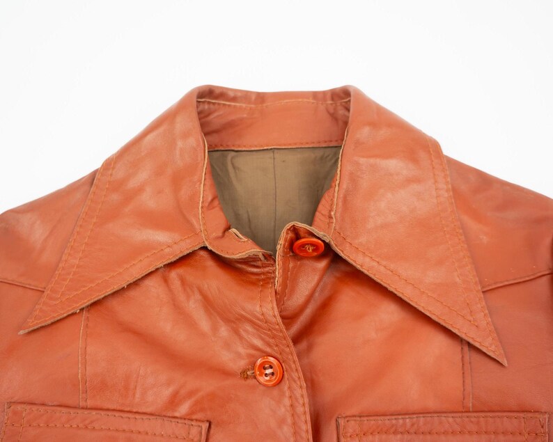 May include: Close-up of a vintage orange leather jacket. The jacket features a wide collar, two front pockets, and orange buttons. The leather has a slightly worn appearance, suggesting its age and use.