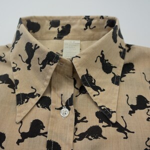 May include: A tan button-up shirt with a black lion print. The shirt has a pointed collar and a single button closure.