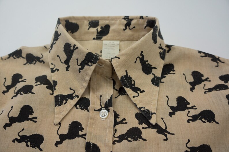 May include: A tan button-up shirt with a black lion print. The shirt has a pointed collar and a single button closure.