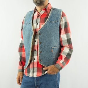 Può includere: Un gilet in denim blu chiaro con fodera color crema e chiusura con bottoni. Il gilet è indossato sopra una camicia a quadri rossa, nera e bianca. Il modello indossa anche jeans blu.
