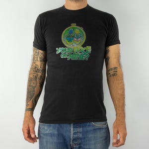 May include: Black t-shirt with a colorful graphic design. The design features a circular emblem with a floral motif and the text "YOUR FACE OR MINE?" in green and white. The shirt is worn by a person.