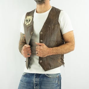 May include: Brown leather vest with a button-up front and side laces. The vest has a small patch on the chest with a skull graphic. The vest is worn over a white t-shirt and blue jeans.