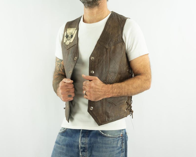May include: Brown leather vest with a button-up front and side laces. The vest has a small patch on the chest with a skull graphic. The vest is worn over a white t-shirt and blue jeans.