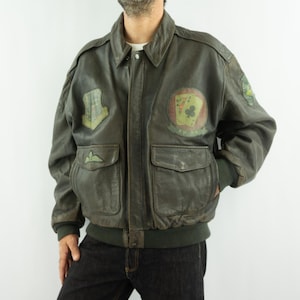 Könnte beinhalten: Vintage olivgrüne Leder-Bomberjacke mit Aufnähern. Die Jacke hat einen klassischen Kragen, zwei Vordertaschen sowie einen gerippten Saum und Bündchen. Zu den Aufnähern gehören ein Spielkarten-Design und militärische Abzeichen.