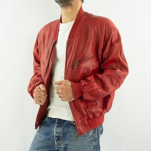 Op de afbeelding: Rode leren bomberjack met ritssluiting en geribde manchetten. De jas heeft een klassiek design met een ritszak op de borst. Het model draagt een blauwe jeans en een wit t-shirt.