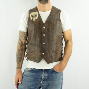 May include: Brown leather vest with a V-neck, button closure, and side lacing. The vest has two front pockets and a small patch on the chest. The vest is worn over a white t-shirt and blue jeans.