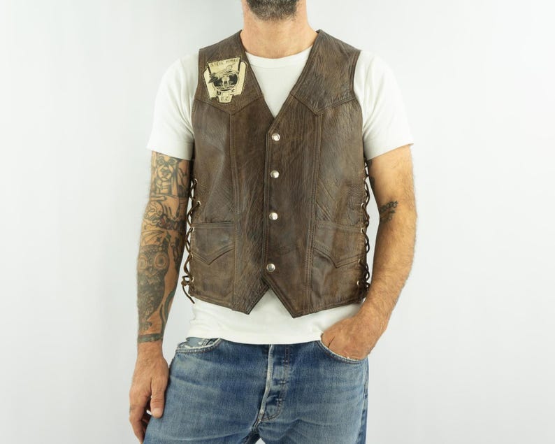 May include: Brown leather vest with a V-neck, button closure, and side lacing. The vest has two front pockets and a small patch on the chest. The vest is worn over a white t-shirt and blue jeans.