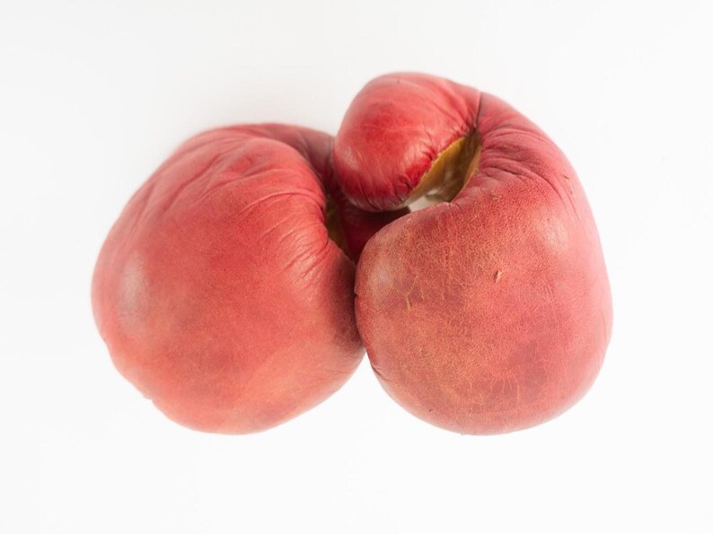 May include: Pair of well-worn, red leather boxing gloves. The gloves are positioned together, with the palms facing inward. The leather shows signs of use, with some discoloration and wear. The gloves are set against a plain white background.