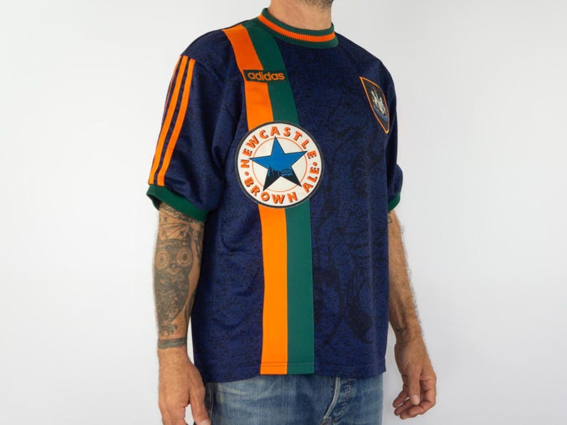 May include: Navy blue Adidas soccer jersey with orange and green accents. Features a Newcastle Brown Ale logo and Adidas logo. The jersey has a round neck and short sleeves.
