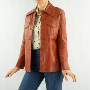 May include: A vintage rust-colored leather jacket with a collared neckline, button-down front, and two chest pockets. The jacket is worn over a patterned button-up shirt and blue jeans. The jacket has long sleeves and a tailored fit.