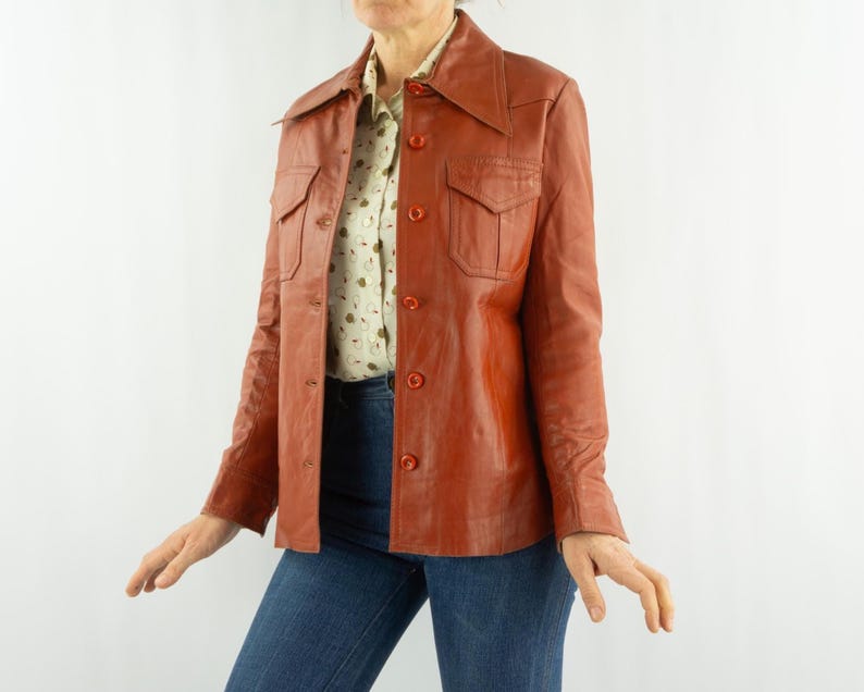 May include: A vintage rust-colored leather jacket with a collared neckline, button-down front, and two chest pockets. The jacket is worn over a patterned button-up shirt and blue jeans. The jacket has long sleeves and a tailored fit.