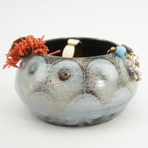 Op de afbeelding: Een keramische kom met een gespikkelde grijze en blauwe glazuur. De kom heeft een donker interieur en is versierd met blauwe en witte cirkelvormige patronen. Koraal en sieraden sieren de rand.