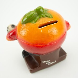 May include: Orange ceramic piggy bank shaped like a grill with a green leaf on top. The piggy bank has a slot on the top and says "Hogar del Pueblo" on the base.