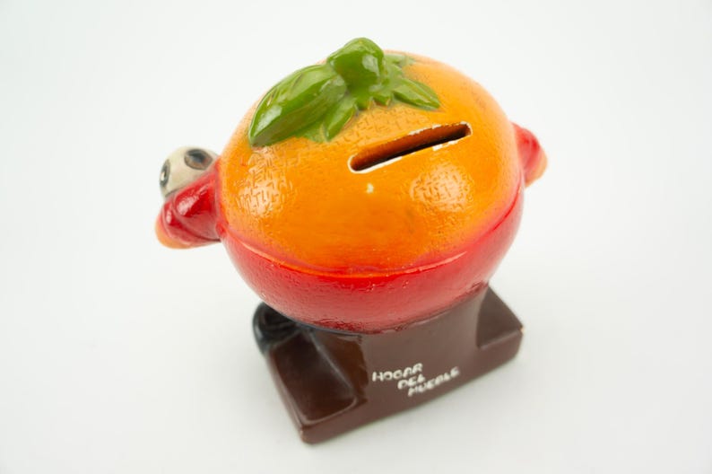 May include: Orange ceramic piggy bank shaped like a grill with a green leaf on top. The piggy bank has a slot on the top and says "Hogar del Pueblo" on the base.