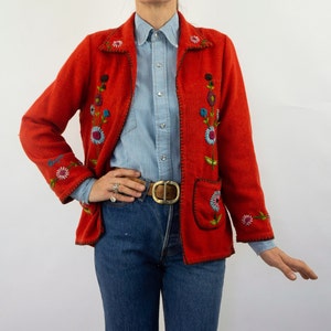 May include: A red embroidered jacket with floral designs. The jacket has a zipper closure and a pocket on the left side. The model is wearing a light blue button-down shirt and blue jeans.