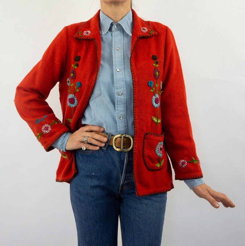 May include: A red embroidered jacket with floral designs. The jacket has a zipper closure and a pocket on the left side. The model is wearing a light blue button-down shirt and blue jeans.
