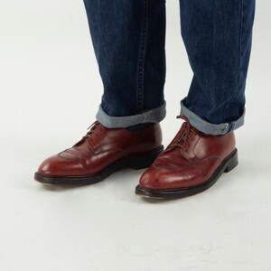 May include: A pair of brown leather lace-up shoes with a pointed toe. The shoes are worn with rolled up blue jeans.