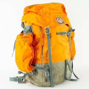 May include: An orange and gray backpack with multiple compartments and adjustable straps. The backpack has a top flap and side pockets. A small patch is visible on the upper right corner. The backpack is designed for outdoor activities.