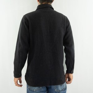 May include: A black wool jacket with a button-down front and long sleeves. The jacket has a collar and buttoned cuffs. The person is wearing blue jeans. The jacket appears to be a medium weight.