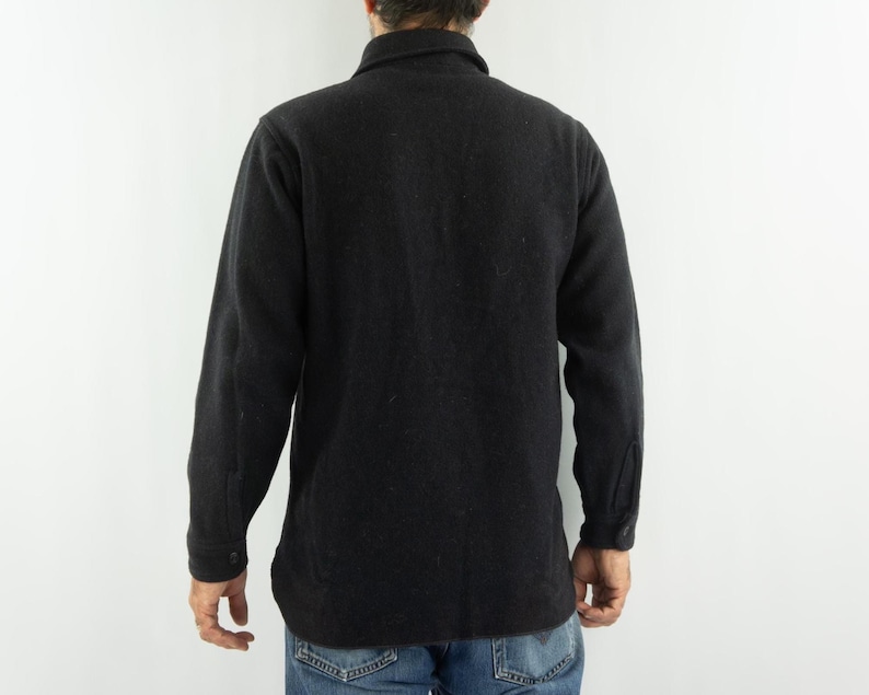 May include: A black wool jacket with a button-down front and long sleeves. The jacket has a collar and buttoned cuffs. The person is wearing blue jeans. The jacket appears to be a medium weight.