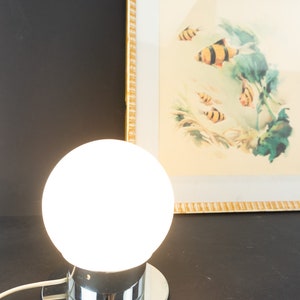 May include: A chrome table lamp with a white glass globe shade. The lamp is turned on and the globe is illuminated.