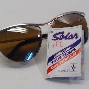May include: Silver ski sunglasses with brown lenses and a tag that reads "Solar Ski collection Tous Temps Guy Perillat France".