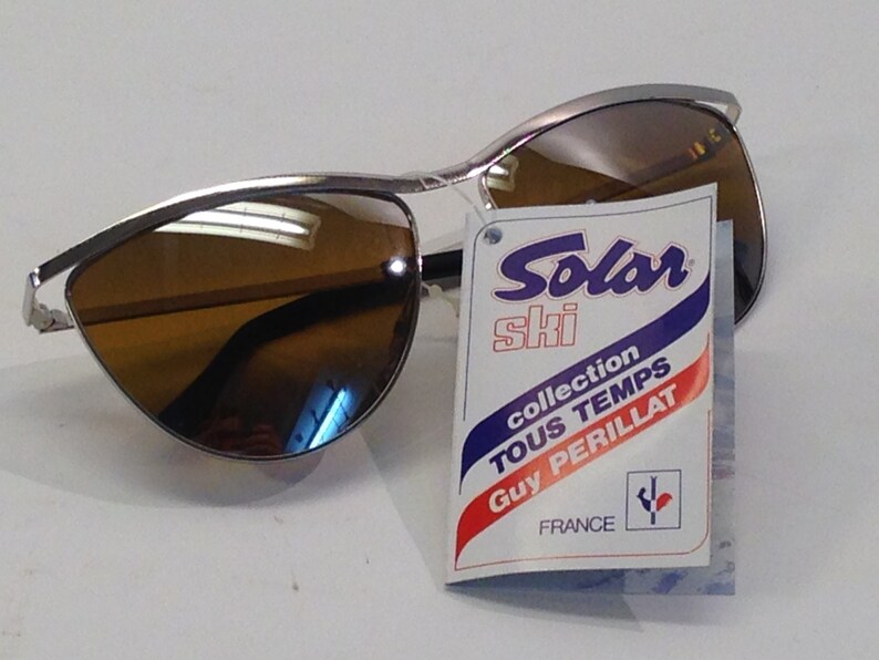 May include: Silver ski sunglasses with brown lenses and a tag that reads "Solar Ski collection Tous Temps Guy Perillat France".