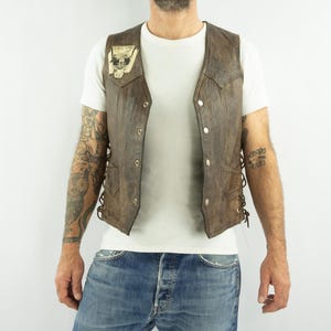May include: Brown leather vest with a skull patch. The vest has a button-up front and lace-up sides. The vest is worn over a white t-shirt and blue jeans.