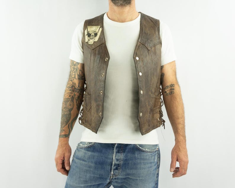 May include: Brown leather vest with a skull patch. The vest has a button-up front and lace-up sides. The vest is worn over a white t-shirt and blue jeans.