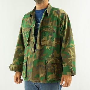 Puede incluir: Una chaqueta de manga larga con botones y un patrón de camuflaje en verde, marrón y negro. La chaqueta incluye dos bolsillos en el pecho con solapa y dos bolsillos delanteros inferiores con solapa, sobre una camiseta azul y vaqueros.