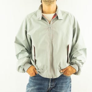 Peut inclure: Une veste zippée gris clair dotée d'un col classique, de manches longues à poignets côtelés et de deux poches latérales. La doublure intérieure présente un motif à carreaux rouge, noir et blanc visible au col et aux poches, portée avec un jean bleu.