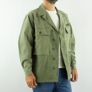 Può includere: Giacca verde oliva con più tasche e chiusure con bottoni. La giacca ha un colletto e una piccola toppa sulla tasca superiore. La persona indossa una maglietta bianca e jeans blu. La giacca è di peso medio.