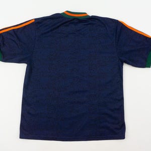 May include: Navy blue short-sleeve shirt with orange stripes on the shoulders and green trim on the sleeves and collar. The shirt has a subtle pattern throughout the fabric.