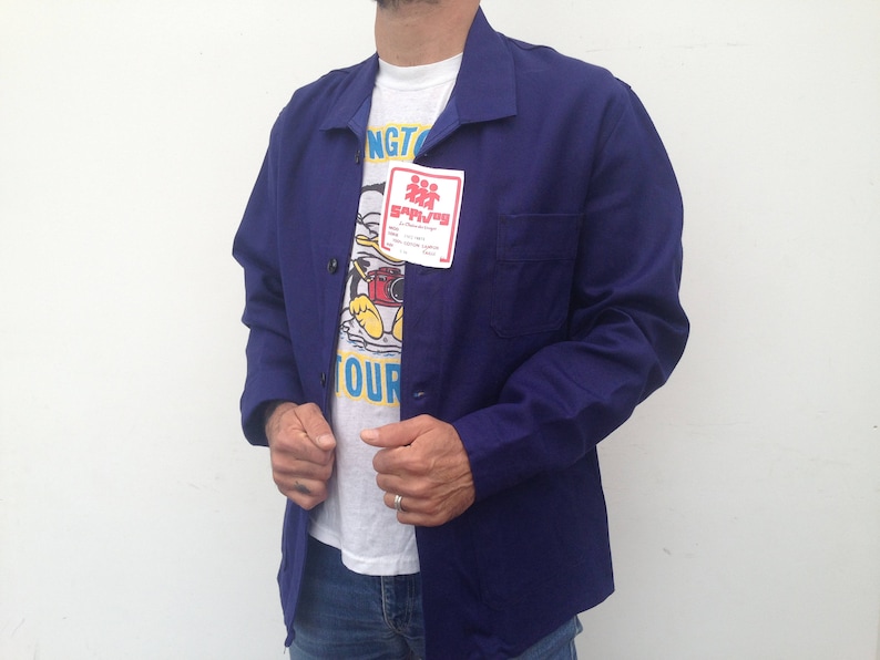May include: A dark blue work jacket with a white t-shirt underneath. The t-shirt has a cartoon character and the text "Sapijoy" on it.
