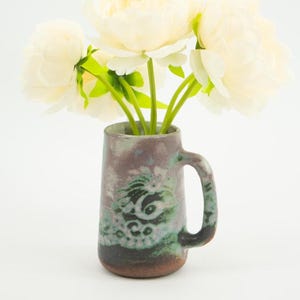 May include: A ceramic mug with a handle, featuring a design in shades of purple, green, and brown. The mug holds a bouquet of white peonies with green stems. The mug is approximately 5 inches tall.