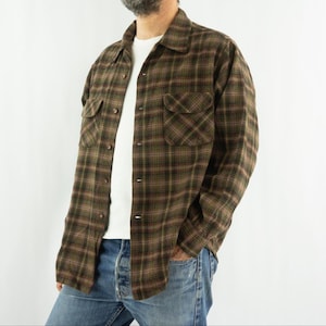 Peut inclure: Une chemise à carreaux marron et verte avec deux poches de poitrine. La chemise est portée sur un t-shirt blanc et un jean bleu. Le motif à carreaux présente des nuances de marron, de vert et de rouge.