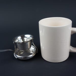 Op de afbeelding: Een witte keramische mok met een gebogen handvat staat naast een zilveren thee-ei. De infuser heeft een kleine komvormige mand en een lange handgreep. De items staan op een donkere achtergrond.