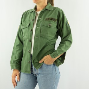 Puede incluir: Chaqueta verde oliva de estilo militar con botones, dos bolsillos en el pecho y cuello. Las palabras "U.S. ARMY" están bordadas en el pecho. La chaqueta se lleva sobre una camisa blanca y combinada con vaqueros azules.