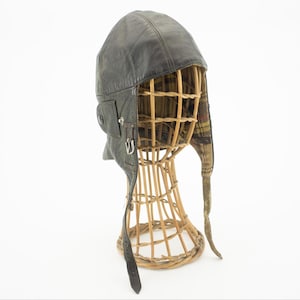 May include: A vintage aviator hat in dark grey leather. It has ear flaps, a chin strap, and a small pocket. The interior lining is a plaid pattern. The hat is displayed on a wicker head form.
